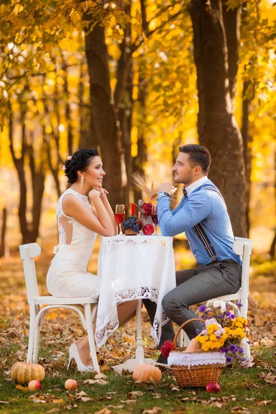 Pareja sonriente sentada en una mesa con decoración en el parque de otoño. Novia y novio felices en el bosque, al aire libre —  Fotos de Stock