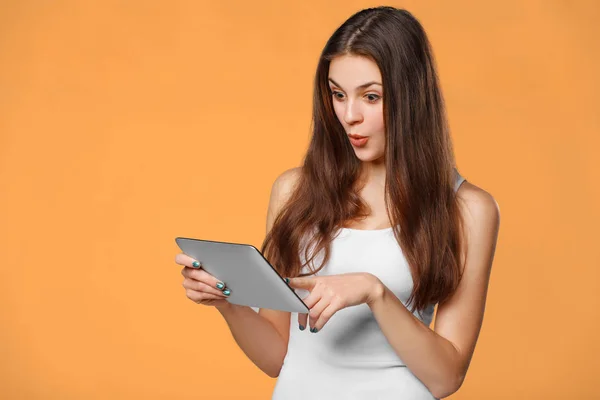 Excited woman with tablet pc. Happy girl in white shirt using tablet, isolated on orange background — Stock Photo, Image