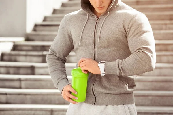 Joven hombre activo va a beber mirando su reloj, horario, al aire libre. Masculino musculoso guapo sosteniendo agitador, agua potable — Foto de Stock