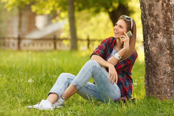 Jovem mulher feliz falando no telefone celular sentado na grama no parque da cidade de verão. Menina moderna bonita com um smartphone, ao ar livre — Fotografia de Stock