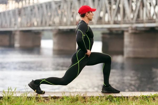 Атлетик делает упражнения на растяжку, на открытом воздухе. Активный мужчина тренируется снаружи на фоне моста — стоковое фото