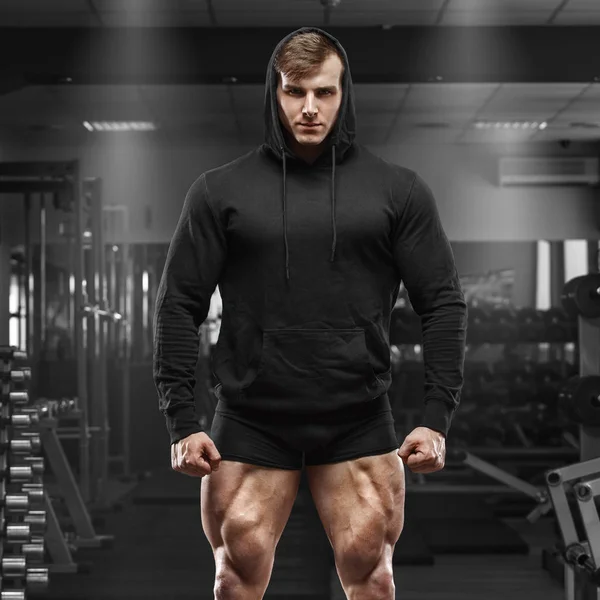 Homem musculoso com pernas musculadas no ginásio. Forte macho em capuz preto com quads grandes — Fotografia de Stock