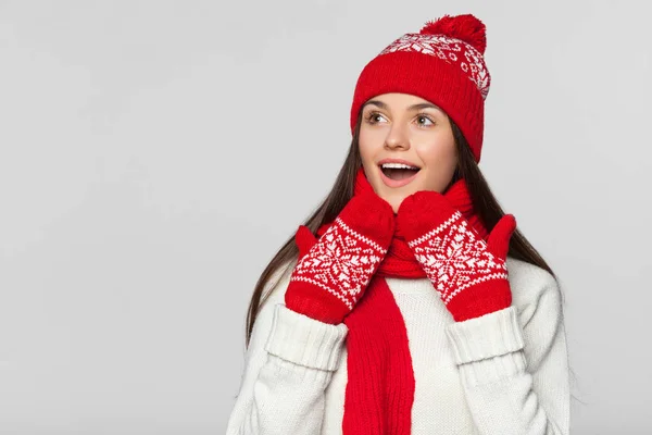 Happy surprised woman looking sideways in excitement. Excited christmas girl wearing knitted warm hat and scarf, isolated on gray background