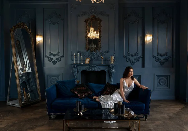 Effektive Brünette in silber glänzendem Kleid posiert auf Sofa im schicken Vintage-Interieur — Stockfoto