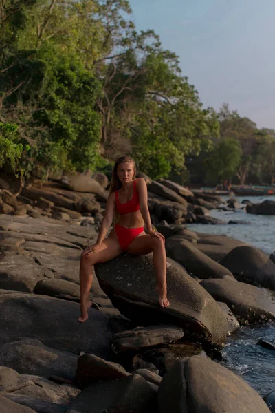 セクシーなめしブルネットで赤ビキニ座っていますザ石の上に熱帯のビーチ — ストック写真