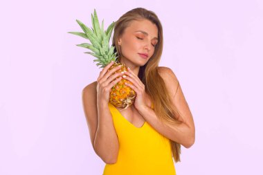 Çekici uzun saçlı esmer kadın portresi. Sarı mayolu. Yüzüne yakın bir ananas tutuyor.