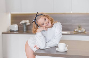 Beyaz kazaklı ve külotlu güzel bir sarışın mutfak masasının yanında fincanla poz veriyor. 