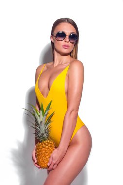 Sarı mayo ve güneş gözlüklü, elinde ananasla beyaz arka planda poz veren seksi, bronz tenli kız.
