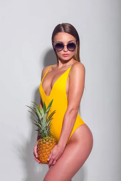 穿着黄色泳衣 戴着墨镜 手上拿着菠萝 背景为白色的性感瘦小女孩 — 图库照片