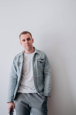 Beyaz tişörtlü ve kot ceketli genç bir adam beyaz bir duvarın karşısında duruyor.