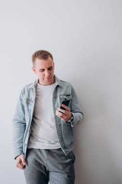 Beyaz tişörtlü ve kot ceketli genç bir adam beyaz bir duvarın karşısında duruyor.