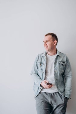 Beyaz tişörtlü ve kot ceketli genç bir adam beyaz bir duvarın karşısında duruyor.