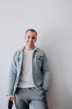 Beyaz tişörtlü ve kot ceketli genç bir adam beyaz bir duvarın karşısında duruyor.