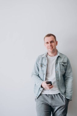 Beyaz tişörtlü ve kot ceketli genç bir adam beyaz bir duvarın karşısında duruyor.