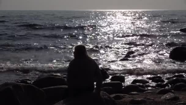 Kvinna siluett sitter stranden. Vatten glittrar i sjö eller hav vid solnedgången. — Stockvideo