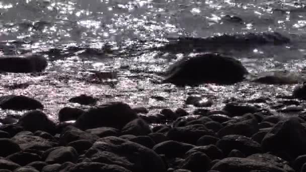 Woda małe fale lśni przez słońce o zachodzie słońca. Jezioro z kamieniami plaży. — Wideo stockowe