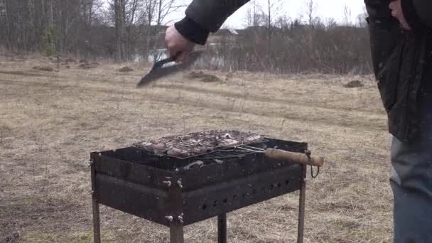 Muž rozdmýchávající oheň na grilu u plastového prkna. s mřížkou plnou masa. — Stock video