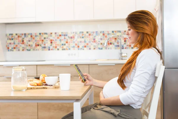 Mujer embarazada preparando comida, verduras, y mirando smartphon — Foto de Stock