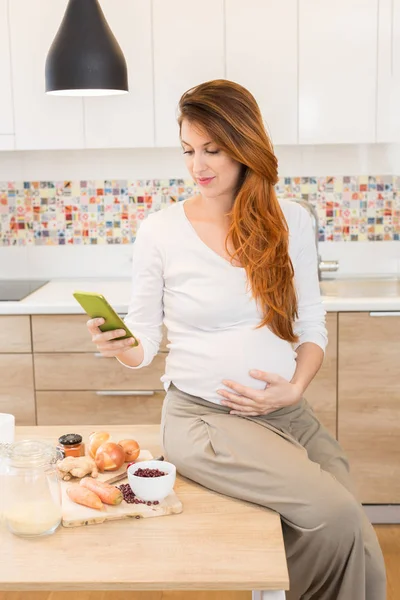 Gravid kvinna förbereder mat, grönsaker, och ser smartphon — Stockfoto