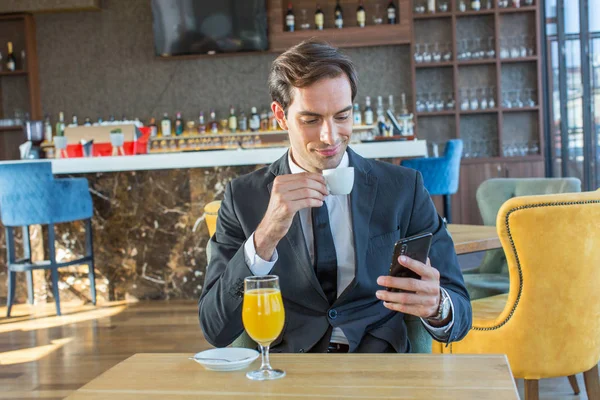 Joven hombre de negocios guapo usando su teléfono celular en la cafetería del hotel —  Fotos de Stock