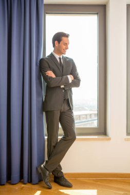 Pencerenin yanındaki otel odasında poz veren yakışıklı, takım elbiseli genç bir işadamı.