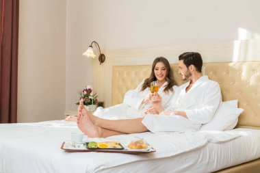 Yatakta kahvaltı eden bornozlu çekici genç bir çift.