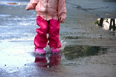 Su geçirmez ceketle su birikintisine atlayan mutlu çocuk. Parlak yağmurluklu bir kız yağmur havasında eğlenir.