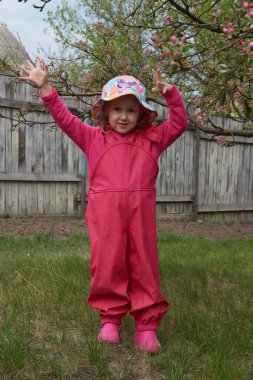 Su geçirmez ceketli ve lastik botlu mutlu çocuk açık havada eğleniyor. Bir kız yağmur havasında kırsalda parlak bir yağmurlukla eğlenir. Bahar çiçeği ağacı.