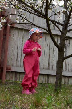 Su geçirmez ceketli ve lastik botlu mutlu çocuk açık havada eğleniyor. Bir kız yağmur havasında kırsalda parlak bir yağmurlukla eğlenir. Bahar çiçeği ağacı.