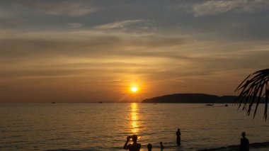 Güzel bir gün batımı bulutlu gökyüzünde parlayan turuncu ışık ile alacakaranlık şafağına dönüşür, denizin üstünde, uzun kuyruk teknesi ve siyah dağ, insanlar akşamları Krabi, Tayland 'da bir sahilde eğlenirler.