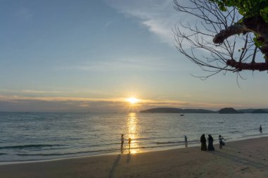 Bulutlu gökyüzünde parlayan turuncu ışıkla güzel bir gün batımı alacakaranlık şafağına dönüşür, denizin üstünde, uzun kuyruk teknesi ve siyah dağ, insanlar akşamları Tayland, Krabi 'de bir sahilde eğlenirler.