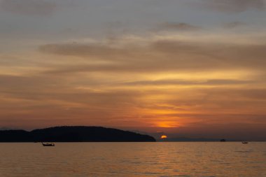Bulutlu gökyüzünde parlayan turuncu gün batımının manzarası akşam vakti Tayland, Krabi 'de bir plajda deniz ve siyah dağ üzerindeki alacakaranlık sahnesine dönüşür.
