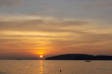Bulutlu gökyüzünde parlayan turuncu ışıkla güzel bir gün batımı alacakaranlık şafağına dönüşür, denizin üstünde, uzun kuyruk teknesi ve siyah dağ, akşam vakti Tayland 'ın Krabi sahilinde.