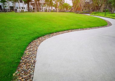 Pürüzsüz yeşil çimenler ve gri eğri desenli yürüyüş yolu. Kum, kahverengi çakıl sınırlı beton kaldırımları yıkıyor. Bakım ve bahçe bakımında destekleyen ve çalı olan ağaçlar.