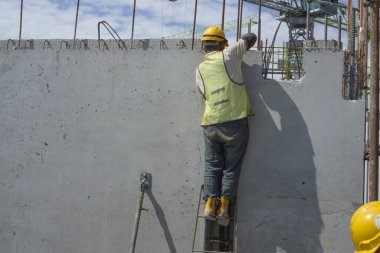 Bir inşaatçı binanın en üst katında çelik çubuklu beton yapı, turuncu güvenlik miğferi ve yeşil yelek, arka planda da bir vinç inşa ediyor.