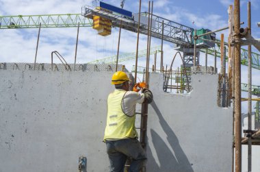 Bir inşaatçı binanın en üst katında çelik çubuklu beton yapı, turuncu güvenlik miğferi ve yeşil yelek, arka planda da bir vinç inşa ediyor.