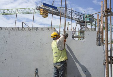 Bir inşaatçı binanın en üst katında çelik çubuklu beton yapı, turuncu güvenlik miğferi ve yeşil yelek, arka planda da bir vinç inşa ediyor.