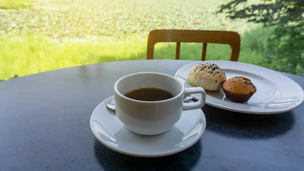 White cup of americano black coffee, cup cake and cereal bread on white plate on black table, green landscape on background under sunshine morning