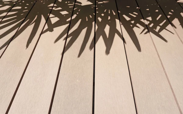 The brown wooden deck and black fade shadow of palm tree leafs, shade on the house\'s terrace from tree\'s leaves in the garden