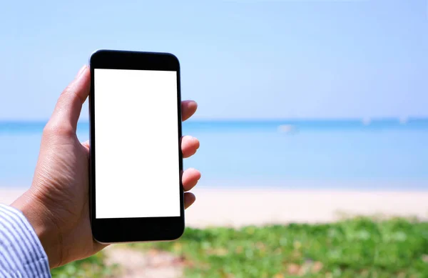A white screen mobile smartphone on a woman\'s hand in front the white sand beach blue sea and sky