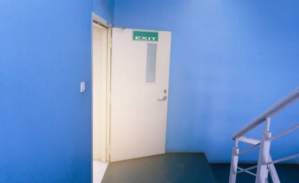 A cream fire exit door opened from a blue wall room to a stair with gray color railing, a green signage with white Exit letters on the top of the door