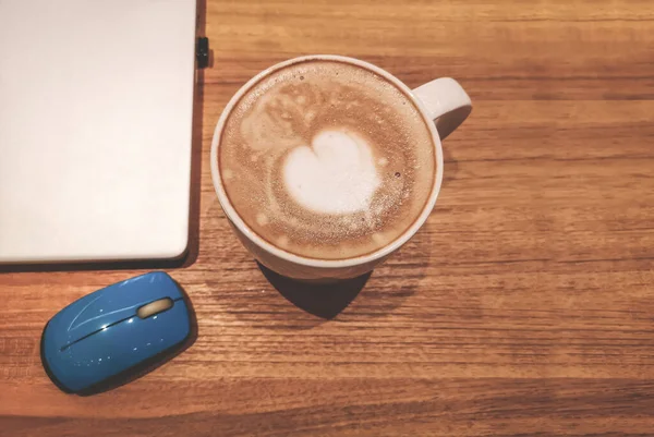 Top view, a white cup hot capuccino with white heart mug on top, a computer labtop and blue mouse on brown wooden table