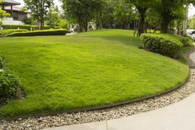 Gri kıvrımlı yürüyüş yolunun bakım bahçesi, beton kaldırımların üzerinde yıkanan kum, kahverengi çakıl sınırı, pürüzsüz yeşil çimenler, destekli ve çalılıklı ağaçlar.