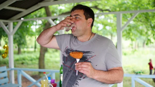 Big man drinking and eating a cutlet in a park pavilion
