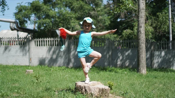 Uma menina está em uma perna em um toco em um parque com uma boneca na mão — Fotografia de Stock