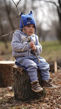 Gri ceketli küçük bir çocuk bahar parkında elinde bir dalla bir kütüğün üzerinde oturuyor.