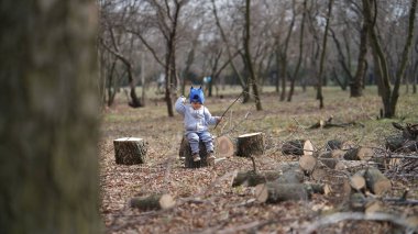 Gri ceketli küçük çocuk bir kütüğün üzerinde oturuyor elinde bir dal, bahar parkında tek başına.