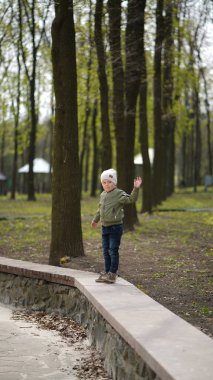 Yeşil ceketli şirin bir çocuk portresi ve baharda parkta beyaz şapka. Çocuk korkulukların üzerinde duruyor ve elini sallıyor.