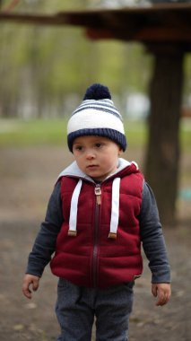 Kırmızı ceketli, baharda parkta beyaz-mavi şapkalı dalgın suratlı küçük şirin bir çocuğun portresi.