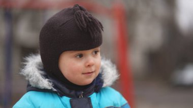 Mavi ceketli, kahverengi şapkalı, çocuk parkında gülümseyen bir çocuğun portresi.